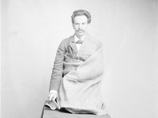 A young man with wavy hair and a bushy moustache, with burlap draped around his body, kneels on a table. A young man with wavy hair and a bushy moustache, with burlap draped around his body, kneels on a table.