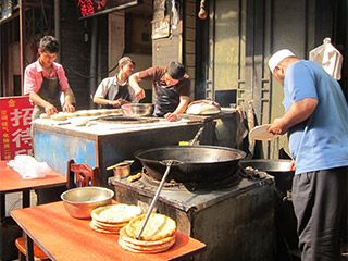 One man stands near a huge pot while three other men prepare food for cooking. One man stands near a huge pot while three other men prepare food for cooking.