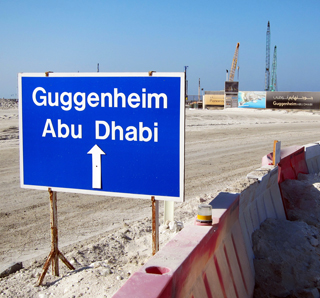 A photograph that features a large blue sign in the foreground that reads "Guggenheim Abu Dhabi." A construction site with cranes can be seen in the background. A photograph that features a large blue sign in the foreground that reads "Guggenheim Abu Dhabi." A construction site with cranes can be seen in the background.