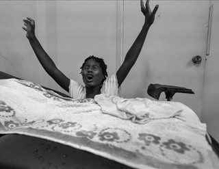 A black woman standing behind a simple hospital bed. Her arms are outstretched, her eyes are close, and her mouth is open, as if she is screaming. A black woman standing behind a simple hospital bed. Her arms are outstretched, her eyes are close, and her mouth is open, as if she is screaming.