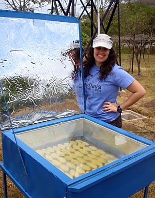 Photo of a student standing outdoors next to a box containing cookies, with clear plexiglass top and a large foil solar reflector. Photo of a student standing outdoors next to a box containing cookies, with clear plexiglass top and a large foil solar reflector.