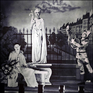 Film still: Onstage in a theater, a live tableau including a mime, a harlequin playing guitar, and Aphrodite in a public garden. Film still: Onstage in a theater, a live tableau including a mime, a harlequin playing guitar, and Aphrodite in a public garden.