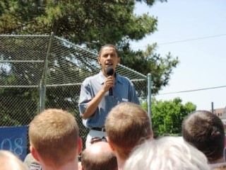 Barack Obama holding a microphone and speaking outdoors in front of a number of people. Barack Obama holding a microphone and speaking outdoors in front of a number of people.