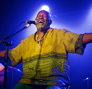 A man in a colorful green and yellow shirt performs on stage. He sits in front of a microphone with arms spread wide and has a hug grin on his face. A man in a colorful green and yellow shirt performs on stage. He sits in front of a microphone with arms spread wide and has a hug grin on his face.