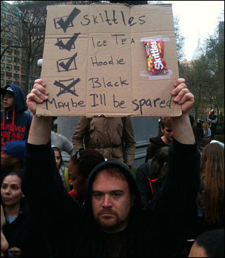A young white man wearing a hoodie holds a sign over his head with the words "Skittles," "Ice tea," and "Hoodie" listed next to boxes with check marks. Beneath them, next to a box with an x marked in it , are the words "Black. Maybe I'll be spared." A young white man wearing a hoodie holds a sign over his head with the words "Skittles," "Ice tea," and "Hoodie" listed next to boxes with check marks. Beneath them, next to a box with an x marked in it , are the words "Black. Maybe I'll be spared."