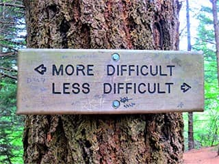A tree with a block of wood fastened to it. Engraved on the block are the words: More Difficult, with an arrow pointing left, and Less Difficult, with an arrow pointing right. A tree with a block of wood fastened to it. Engraved on the block are the words: More Difficult, with an arrow pointing left, and Less Difficult, with an arrow pointing right.
