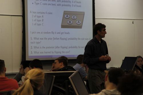 <p>During each class, students worked on two to three board problems, each with multiple parts. We found that three cycles of getting up, working, and sitting down was the limit of student energy. Beyond that they would drag their feet.</p> <p>Problem statements were projected on screens around the room.</p> n instructor stands amidst seated students. The screen on the wall shows a slide with a math problem for the students to work on.