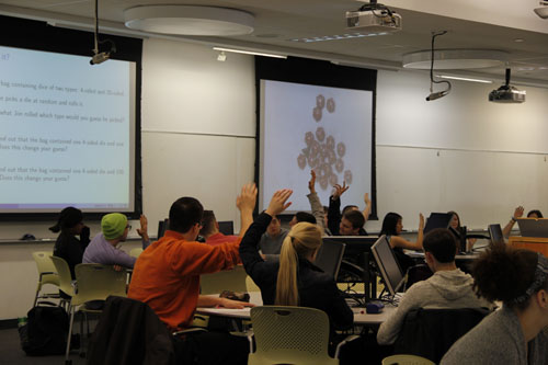 <p>In many class meetings, we illustrated concepts using physical objects like dice and cards. Sometimes, this took the form of an instructor-led demonstration in front of all of the students. We often asked students to participate by choosing cards, guessing outcomes, and explaining their ideas.</p> <p>At the start of the course, we also gave each student a bag of dice that included 10-sided dice and all the Platonic solids. The students used the dice to explore probability and collect data for statistics.</p> <p>In the demonstration pictured here, we illustrated the role of prior probability in Bayesian inference. We told students that we would randomly select and roll a die from a cup containing dice with 4-sides and 20-sides. Secretly the cup contained twenty 20-sided dice and one 4-sided die, and we intentionally placed the 4-sided die on top of the pile in order to select it by feel. We rolled the die covertly, reported a 3, and asked students which type of die was most likely chosen; students were split between 4-sided and “equally likely”. We then used the document camera to reveal the contents of the cup; as shown, the students now overwhelmingly favored the 20-sided die. This demonstration can be seen in the sample class video.</p> Students seated at round tables raise their hands. One projector screen shows a slide and one projector screen shows a set of dice.