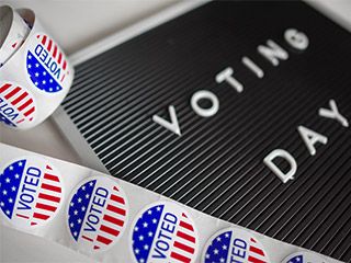 A strip of red, white, and blue stickers with the words “I Voted” lies next to an announcement board with the words “Voting Day” written upon it. A strip of red, white, and blue stickers with the words “I Voted” lies next to an announcement board with the words “Voting Day” written upon it.
