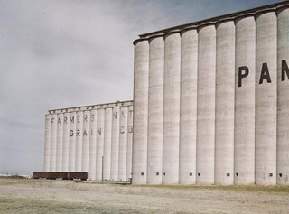 A photo of two rows of tall concrete grain elevators with a few railroad cars. A photo of two rows of tall concrete grain elevators with a few railroad cars.