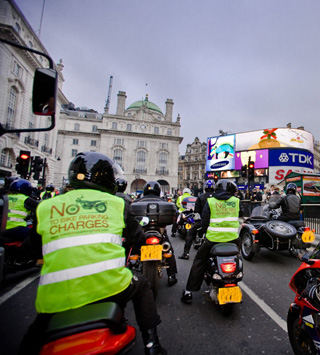 Photo of a large group of motorcyclists riding down a city street, some wearing vests with protest message 'No to bike parking charges!' Photo of a large group of motorcyclists riding down a city street, some wearing vests with protest message 'No to bike parking charges!'