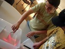Two students stand at a table where there is a plastic box with small laser beams reflected inside.